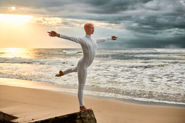 Young hairless ballerina with alopecia in white futuristic suit dancing on seashore at sunset sea,...