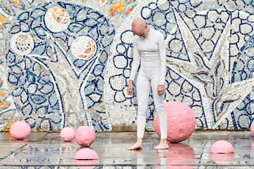 Young hairless girl ballerina with alopecia in white futuristic suit stands outdoor among pink...