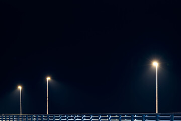 Street lampposts casts dim cold blue glow along shore pier standing against pitch black canvas of...