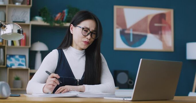 Asian freelancer having online meeting on laptop with her business partner or customer. Young girl in glasses for better vision actively talking. Writing important information on notebook.