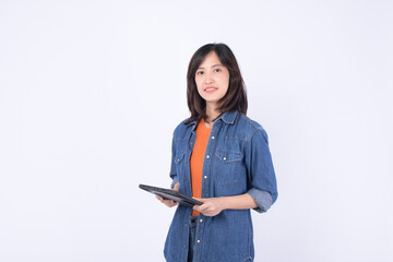 Asian woman wearing orange shirt and denim jean jacket is holding a tablet against a white...