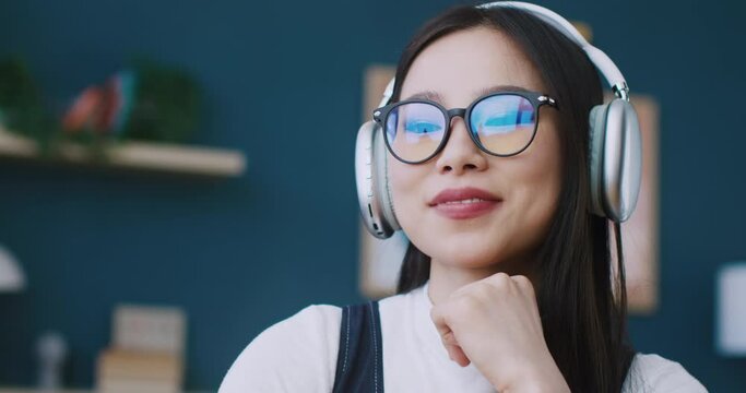 Beautiful Asian woman in glasses putting headphones on her head. Listening music on digital gadget. Enjoying favourite trek or playlist. Concept of leisure at home. Relaxed atmosphere.