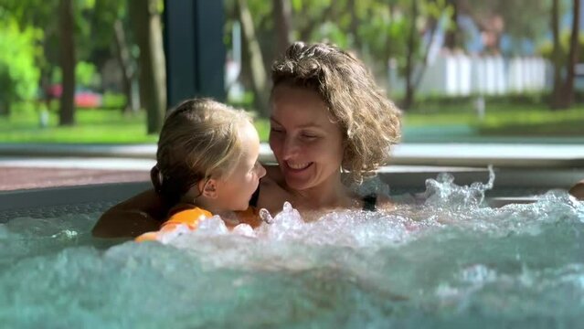 Mom and daughter have fun at spa, splashing around in jacuzzi. Mother child funny splash with water and truly enjoy time spent in jacuzzi. Foam bubbles warm jacuzzi water charge with positive energy.