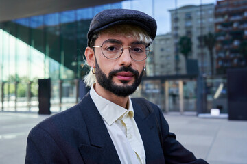 Portrait of a young hipster businessman looking at camera with a serious and confident face, wearing a beret hat, standing outside financial district.