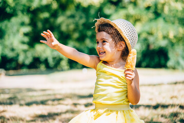 Cute happy little girl in yellow dress and hat eating fruit ice cream. Summer fun child playing in park. Summertime vibrant color background.