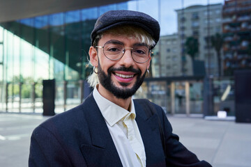 Portrait of a young hipster businessman looking at camera smiling standing outside workplace. Caucasian male wearing a beret and a suit.