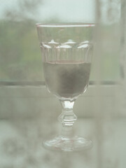 Vertical close up view of a glass of red wine on a windowsill behind a white laced curtain.