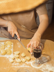 A vertical close-up captures a woman making dumplings, her face obscured by a straw lampshade, as she skillfully cuts dough with a knife. A glass of red wine sits on the messy table nearby.