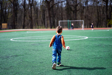 A young boy in a pair of overalls is walking across a green field with a soccer ball in his hand