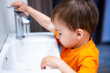 A young boy is playing with the water in a sink. He is wearing an orange shirt. The sink is white...