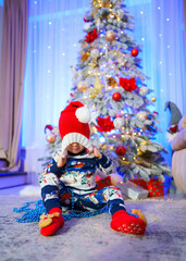 Fototapeta premium A young child is sitting in front of a Christmas tree, wearing a red hat and a blue and white snowsuit