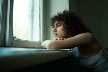 The girl with curls is leaning on the windowsill and looking out the window. Grain effect used