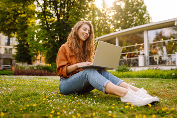 Portrait of attractive young woman sitting on green grass in park while using laptop and wireless earphone. Freelancer, education, online, technology concept.