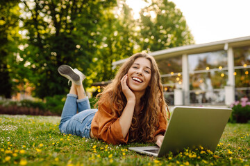 Portrait of attractive young woman sitting on green grass in park while using laptop and wireless...