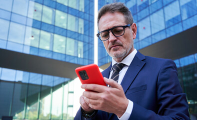Senior businessman using phone to work, standing outside of a corporate district, concentrated on the device, sending online messages to be connected. Copy space.