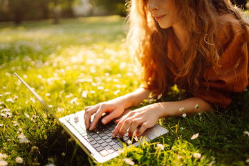 Portrait of attractive young woman sitting on green grass in park while using laptop and wireless...