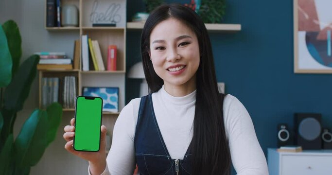 Positive Asian girl holding phone with green screen in her hands. Cute smiling and looking at camera. Holding up big finger while recommending smartphone with chroma key for sale or use.