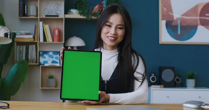 Chinese woman holding tablet device while sitting at table in modern room. Cheerfully smiling and looking directly at camera. Green screen of digital gadget. Chromakey. Showing new model of technology