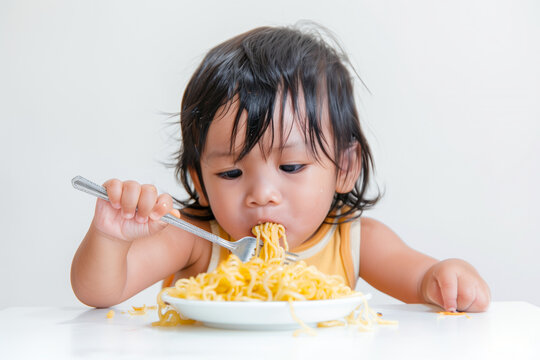 crisp white background, a rich Indonesian toddler delights in slurping a string of fried noodles using a fork, showcasing the joy of indulging in delicious cuisine from an early ag
