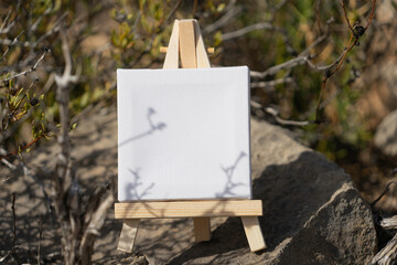 Mockup. White mini canvas on small wooden easel stand standing on cliff rocks outdoors with blurred...