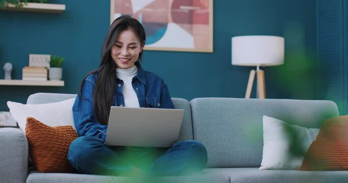 Charming Asian woman sitting in lotus position on large sofa and working from home network on laptop. Pretty girl using technology device for remote job or study. Technology concept.