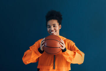 Happy basketball player with a ball over the blue backdrop. Smiling woman with a basketball looking...