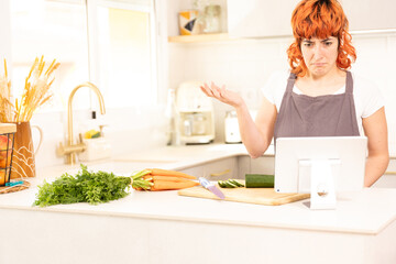 A woman is standing in a kitchen with a tablet in front of her. She is looking at the screen and she is confused. The kitchen is filled with various items such as carrots, broccoli, and a knife