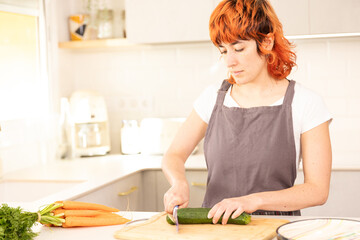 A woman is cutting a cucumber on a cutting board. She is wearing a grey apron and has red hair
