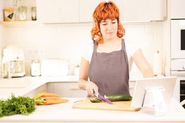 A woman is cutting vegetables on a cutting board while looking at a tablet. She is wearing an apron...