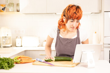 A woman is cutting vegetables on a cutting board in a kitchen. She is using a tablet to look up a recipe