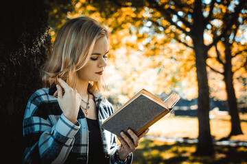 Girl in the jacket attentively reading book on leans on the tree and twirls a curl with her finger