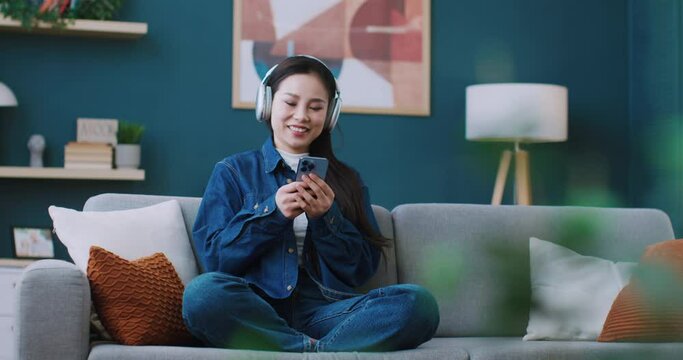 Chinese girl in jeans costume comfortably sitting in lotus position on soft couch in living room. Positive woman listening music in headphones while remotely communicating in social media from phone.