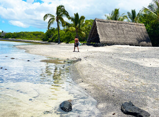 Surfer walking by traditional thatched grass hut hale on tropical island of Hawaii, lagoon, coco palms, blue sky, sandy beach, leisurely stroll, calming, paradise, Hawaiian