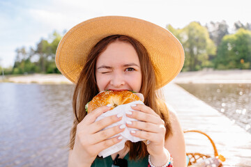 A happy woman in nature wearing a straw hat is enjoying eating a bagel