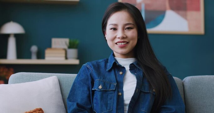 Pretty Asian woman in jeans jacket comfortably sitting on soft couch in living room. Smiling with joy while looking directly at camera. Peaceful leisure at home. Concept of recreation.