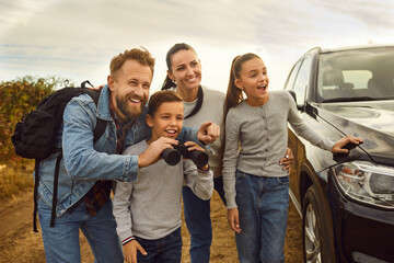 Family travelling by car enjoying recreation camping weekend day outside. Dad with backpack and binoculars looks with mum and kids at countryside landscape view and points finger at animal in distance