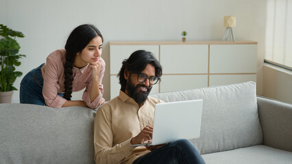 Arabian Indian couple on couch use laptop computer shopping online together pay bills order food at...