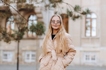 A modern young woman with sunglasses on her eyes which looks at the camera is wearing a black sweater and a beige raincoat and stands against the background of a beautiful grey building