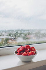 White bowl of organic fresh strawberries on a windowsill with raindrops. Fresh strawberries in the...