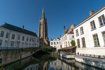 Wharf on the canal since Dijver park with the Our Lady church tower reflected on the canal in the old town of the beautiful city of Brugge in Belgium in a sunny day.