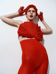 Elegant woman in red dress and gloves posing with hands on head against white wall