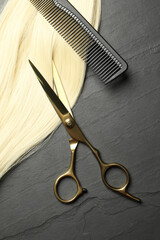 Professional hairdresser scissors and comb with blonde hair strand on dark grey table, flat lay