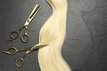 Professional hairdresser scissors with blonde hair strand on dark grey table, flat lay. Space for...