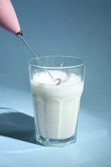 Whisking milk in glass with mini mixer (milk frother) on light blue background