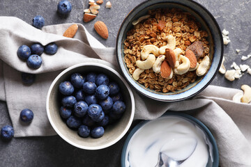 Tasty granola in bowl, blueberries, yogurt and spoon on gray textured table, flat lay