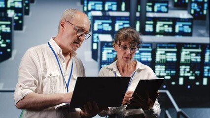 Fototapeta premium Knowledgeable IT practitioners looking around server farm, using laptop and tablet to crosscheck disaster recovery plan and assess rackmounts in need of replacement, preventing perils