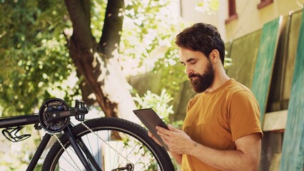 Determined active man examining and fixing his bicycle while using his digital tablet for guidance....