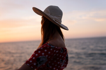 A woman wearing a straw hat is sitting on a rock near the ocean.