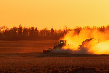 Silhouette of a tractor seeding in dust in the field in the orange sunset.