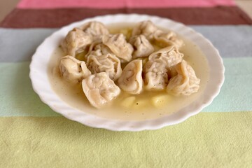 Soup with beef dumplings and potatoes in a white plate on a multi colored tablecloth on a table at home. It is steaming. Close-up